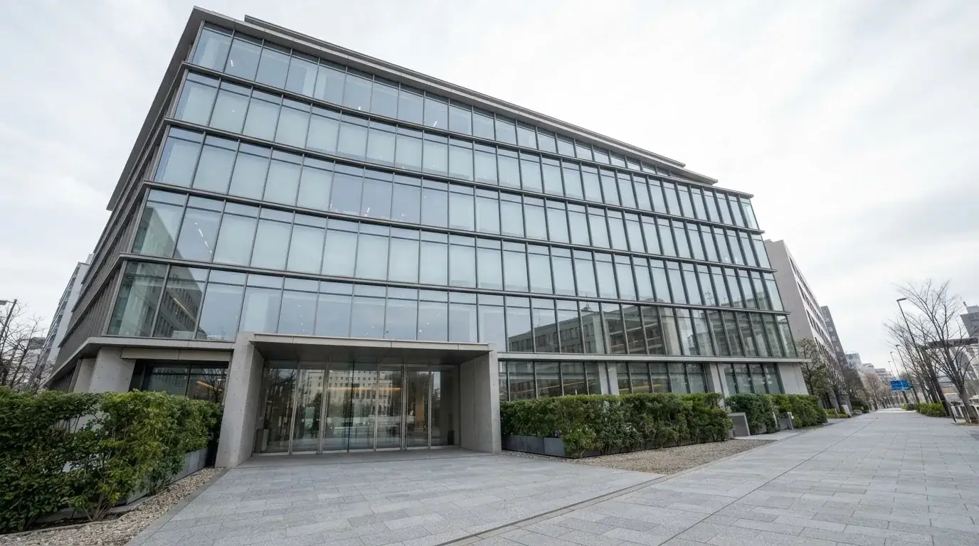 Edificio moderno de oficinas con grandes ventanales de cristal reflejando el cielo, visto desde una calle peatonal.